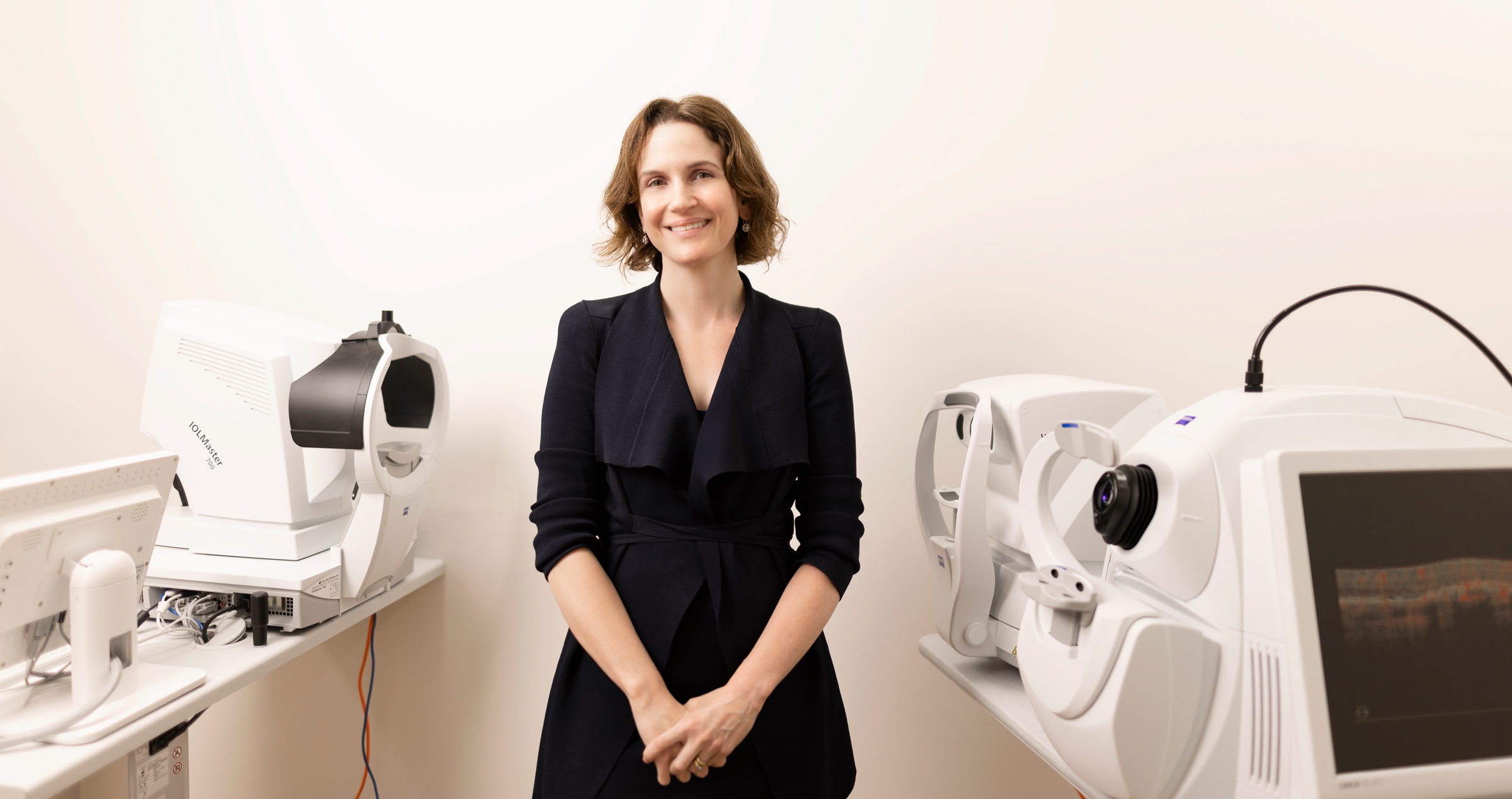 Dr Madeleine Adams wearing a black dress, smiling and standing in the clinic surrounded by ophthalmologist equipment